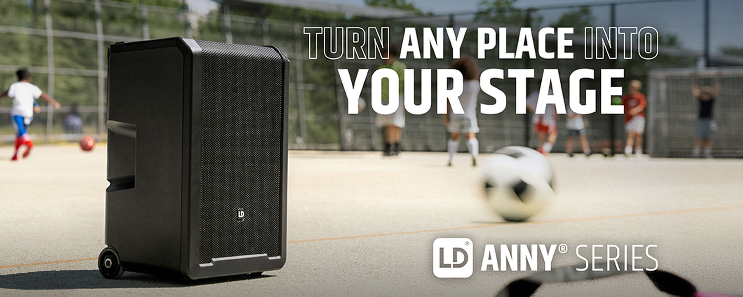 Mobiler Lautsprecher der LD Systems ANNY Serie auf einem Sportplatz mit Fußball spielenden Kindern im Hintergrund und dem Slogan „Turn any place into your stage“